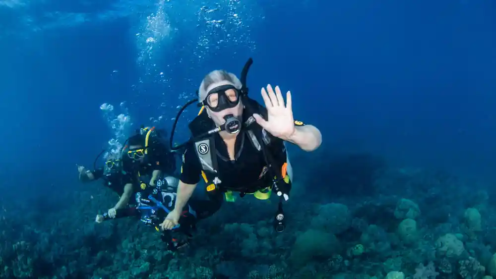 A diver waving to the camera above a coral slope